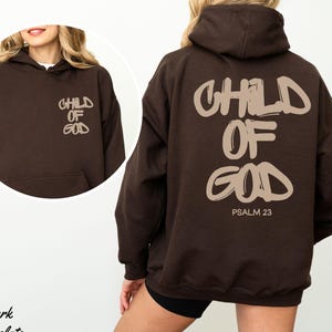 Pode incluir: Moletom marrom escuro com o texto "CHILD OF GOD" em uma fonte estilizada marrom claro na frente e nas costas. A parte de trás também inclui "PSALM 23". O moletom tem um bolso frontal e um capuz.