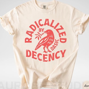 May include: Ivory-colored t-shirt with rolled sleeves. The shirt features a red graphic of a bird holding a branch, surrounded by the words "RADICALIZED BY BASIC DECENCY" in a circular design. The shirt is made of a soft material.