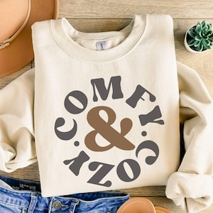 May include: A sand-coloured sweatshirt with the words "COMFY & COZY" in a circular design. The text is in shades of grey and brown. The sweatshirt is folded on a wooden surface, with a hat and jeans visible.