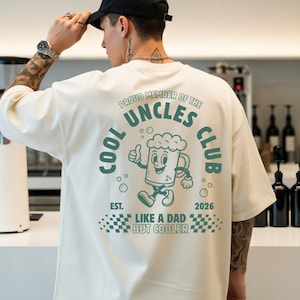 May include: Ivory-colored t-shirt with a graphic on the back that reads "COOL UNCLES CLUB" with a cartoon beer mug character. The shirt also has the text "LIKE A DAD BUT COOLER" and "2026". The brand "COMFORT COLORS" is visible.