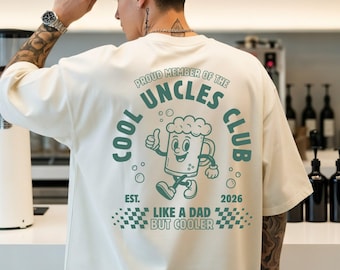 Può includere: T-shirt color avorio con grafica sul retro con la scritta "COOL UNCLES CLUB" con un personaggio a fumetti di boccale di birra. La maglietta ha anche il testo "LIKE A DAD BUT COOLER" e "2026". Il marchio "COMFORT COLORS" è visibile.