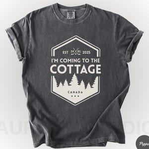 May include: Dark gray t-shirt with rolled-up sleeves. The shirt features a white graphic with the text "I'M COMING TO THE COTTAGE" and "CANADA". The graphic includes a forest silhouette and the year "2025".