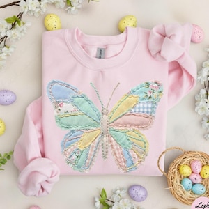 Può includere: Felpa rosa chiaro con un'applicazione a farfalla colorata. Il design della farfalla incorpora vari tessuti pastello, tra cui motivi floreali e a quadretti. L'immagine include uova di Pasqua decorative e accenti floreali, con il testo "Light Pink".