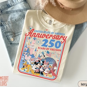May include: Ivory-colored t-shirt with a graphic celebrating the 250th anniversary of the United States. The design features cartoon characters, fireworks, and the text "United State of America Anniversary 250 Years of Freedom". The shirt is paired with light blue jeans and a tan hat.