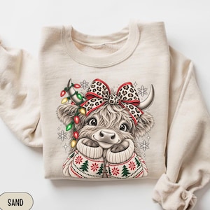Puede incluir: Una sudadera de cuello redondo color arena con el gráfico de una vaca highland peluda que lleva un suéter estampado con árboles verdes y detalles rojos. La vaca tiene un lazo de leopardo, luces navideñas y copos de nieve alrededor.