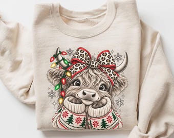 May include: A sand-coloured crewneck sweatshirt featuring a graphic of a fluffy highland cow wearing a patterned jumper with green trees and red accents. The cow has a leopard print bow, string lights, and snowflakes around its head.