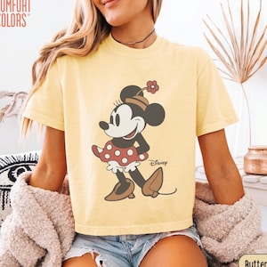 May include: Butter yellow Comfort Colors t-shirt featuring a Minnie Mouse graphic. The cartoon character is depicted in a red polka dot dress, brown shoes, and a matching hat with a flower. The Disney logo is printed on the shirt.