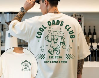Può includere: T-shirt color avorio con grafica verde sul retro con la scritta "COOL DADS CLUB" e un boccale di birra a fumetti. Il testo "EST. 2026" e i nomi "LIAM & EMILY & NOAH" sono stampati sulla maglietta.