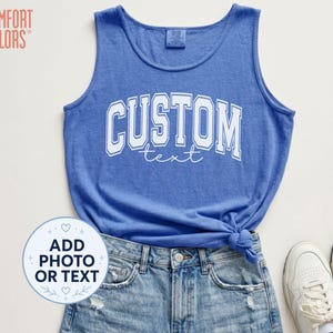 May include: A blue sleeveless tank top featuring the white text CUSTOM text in a collegiate font. The garment has a round neckline and is paired with light blue distressed denim shorts and white sneakers. A circular graphic reads ADD PHOTO OR TEXT.