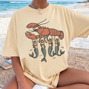 Puede incluir: Una camiseta de manga corta color mantequilla con una ilustración gráfica de una langosta naranja grande sostenida por cuatro sirenas con colas verdes. El diseño está centrado en el pecho de la camiseta, que tiene cuello redondo.