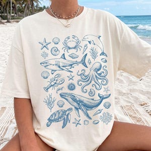 Pode incluir: Uma t-shirt de manga curta na cor marfim com uma estampa gr&aacute;fica azul de v&aacute;rias criaturas marinhas, incluindo uma baleia, um tubar&atilde;o, um polvo, uma tartaruga marinha, um golfinho, um caranguejo, uma lagosta, estrelas-do-mar e conchas.