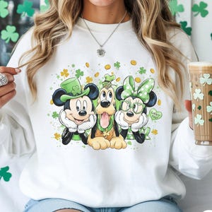 May include: White sweatshirt featuring Mickey Mouse, Goofy, and Minnie Mouse in St. Patrick's Day attire. Mickey wears a green top hat, Goofy has a green shamrock, and Minnie has a green bow and glasses. The phrase "Lucky You!" is on a green heart.
