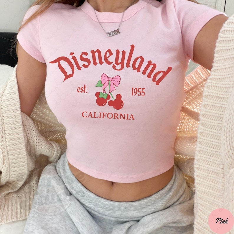 Puede incluir: Top corto rosa con la palabra "Disneyland" en escritura roja, junto con "est. 1955" y "CALIFORNIA". El dise&ntilde;o incluye un gr&aacute;fico de cerezas con un lazo rosa. La camiseta tiene mangas cortas.