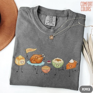 May include: A charcoal gray crew neck t-shirt with a Comfort Colors label at the inner collar. The graphic features cartoon-style Thanksgiving food items including a roll with a flag reading Thankful, a turkey, stuffing, mashed potatoes, and pie.