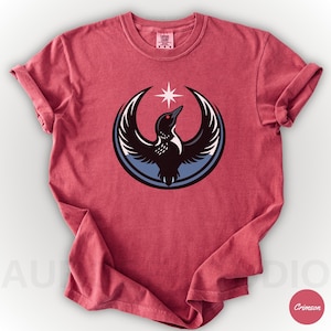 K&ouml;nnte beinhalten: Karmesinrotes T-Shirt mit einer Grafik eines Vogels mit ausgebreiteten Fl&uuml;geln in einem blau-wei&szlig;en Kreis, gekr&ouml;nt von einem Stern. Der Vogel ist schwarz mit wei&szlig;en Akzenten und einem roten Auge. Das Shirt hat einen Rundhalsausschnitt und hochgekrempelte &Auml;rmel.