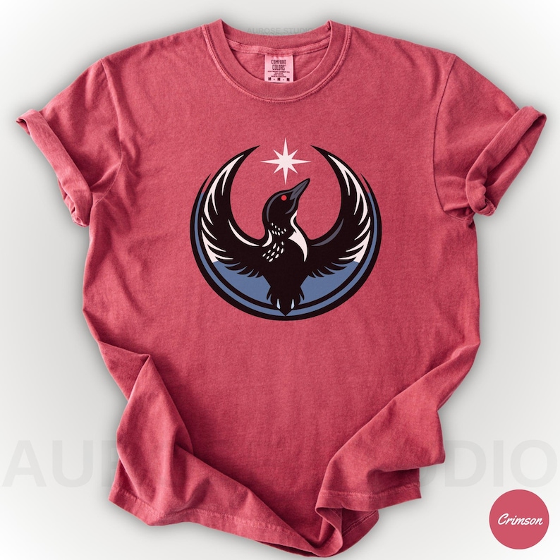 K&ouml;nnte beinhalten: Karmesinrotes T-Shirt mit einer Grafik eines Vogels mit ausgebreiteten Fl&uuml;geln in einem blau-wei&szlig;en Kreis, gekr&ouml;nt von einem Stern. Der Vogel ist schwarz mit wei&szlig;en Akzenten und einem roten Auge. Das Shirt hat einen Rundhalsausschnitt und hochgekrempelte &Auml;rmel.