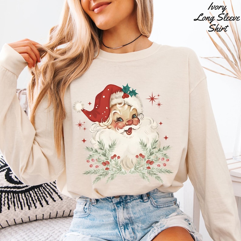 May include: Ivory long-sleeve shirt featuring a vintage Santa Claus graphic. The Santa illustration has a red hat, white beard, and is surrounded by greenery and berries. The shirt has a crew neck and is made of a soft material.