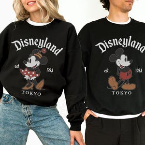 Puede incluir: Sudaderas negras con ribete blanco con el logotipo de Disneyland y gr&aacute;ficos de Mickey y Minnie Mouse. Las sudaderas tienen el texto "est. 1983 TOKYO". Llevadas por dos personas.