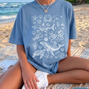 Pode incluir: Uma t-shirt azul de ganga com uma impress&atilde;o gr&aacute;fica branca de v&aacute;rias criaturas marinhas, incluindo um tubar&atilde;o, baleia, polvo, tartaruga marinha, caranguejo, lagosta, golfinho, estrelas-do-mar e v&aacute;rias conchas num padr&atilde;o central.