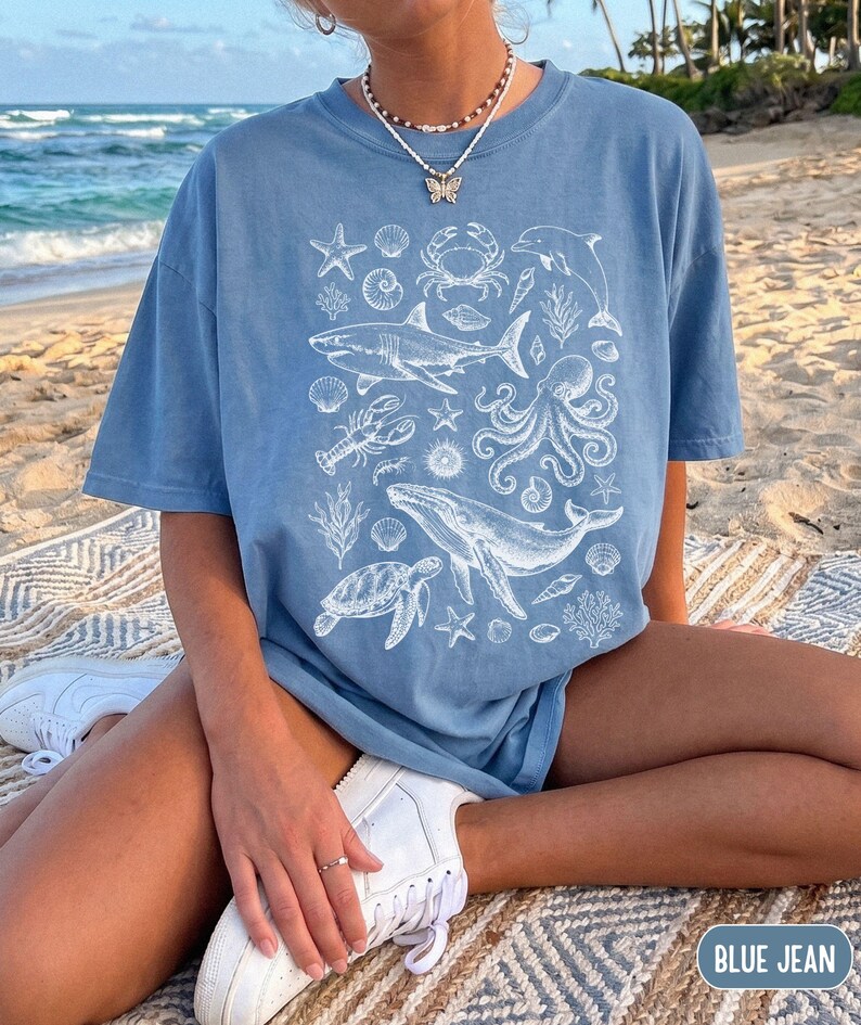 Pode incluir: Uma t-shirt azul de ganga com uma impress&atilde;o gr&aacute;fica branca de v&aacute;rias criaturas marinhas, incluindo um tubar&atilde;o, baleia, polvo, tartaruga marinha, caranguejo, lagosta, golfinho, estrelas-do-mar e v&aacute;rias conchas num padr&atilde;o central.