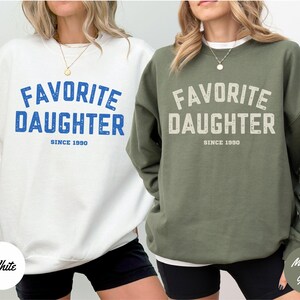 K&ouml;nnte beinhalten: Zwei Sweatshirts, eines in Wei&szlig; und eines in Milit&auml;rgr&uuml;n, mit dem Text "FAVORITE DAUGHTER SINCE 1990" in fetter Schrift. Das wei&szlig;e Sweatshirt hat blauen Text, das gr&uuml;ne hat cremefarbenen Text.