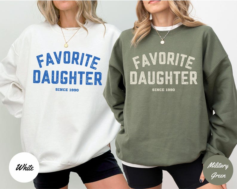 K&ouml;nnte beinhalten: Zwei Sweatshirts, eines in Wei&szlig; und eines in Milit&auml;rgr&uuml;n, mit dem Text "FAVORITE DAUGHTER SINCE 1990" in fetter Schrift. Das wei&szlig;e Sweatshirt hat blauen Text, das gr&uuml;ne hat cremefarbenen Text.