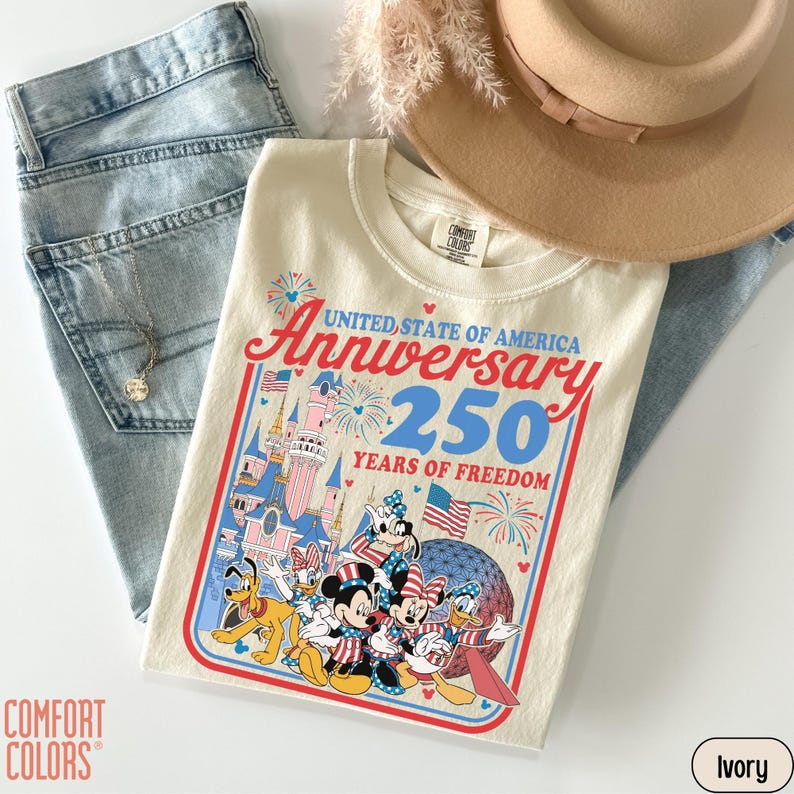 May include: Ivory-colored t-shirt with a graphic celebrating the 250th anniversary of the United States. The design features cartoon characters, fireworks, and the text "United State of America Anniversary 250 Years of Freedom". The shirt is paired with light blue jeans and a tan hat.