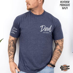 Może przedstawiać: Granatowa koszulka z napisem "Dad" w białym skrypcie i tekstem "EST.2025". Lewy rękaw ma imiona "Liam, Emma, John" w kolorze białym. Koszulka jest z Aurose Studio.