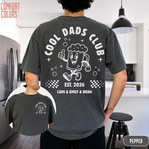 以下が含まれることがあります： 背中に白いグラフィックが入ったペッパーカラーのTシャツ。「COOL DADS CLUB」の文字と、親指を立てた漫画のビールジョッキが描かれています。「EST. 2026」の文字と「LIAM & EMILY & NOAH」の名前もプリントされています。