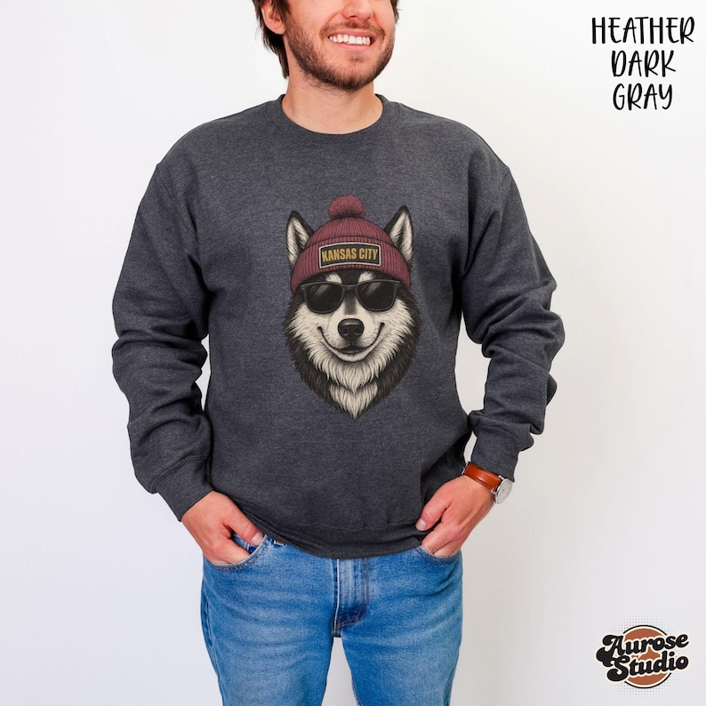 Op de afbeelding: Donkergrijze sweatshirt met een afbeelding van een husky met een zonnebril en een bordeauxrode beanie met "Kansas City" erop. De sweatshirt is heather donkergrijs van kleur.