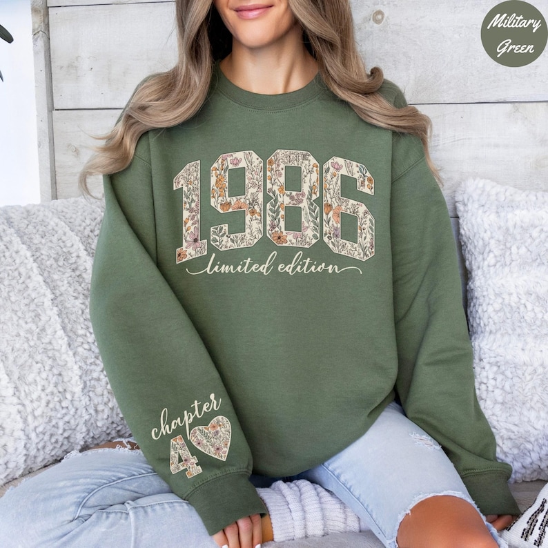 May include: A military green sweatshirt with the year "1986" in a floral design. The words "limited edition" are below the numbers. The left sleeve has the words "chapter 40" with a floral heart design.