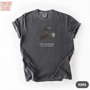 May include: A charcoal gray short-sleeve t-shirt featuring a graphic of a black crow holding a small white mug. Below the crow, the text reads HELLO DARKNESS MY OLD FRIEND. The shirt has a crew neckline and a vintage-style washed fabric finish.