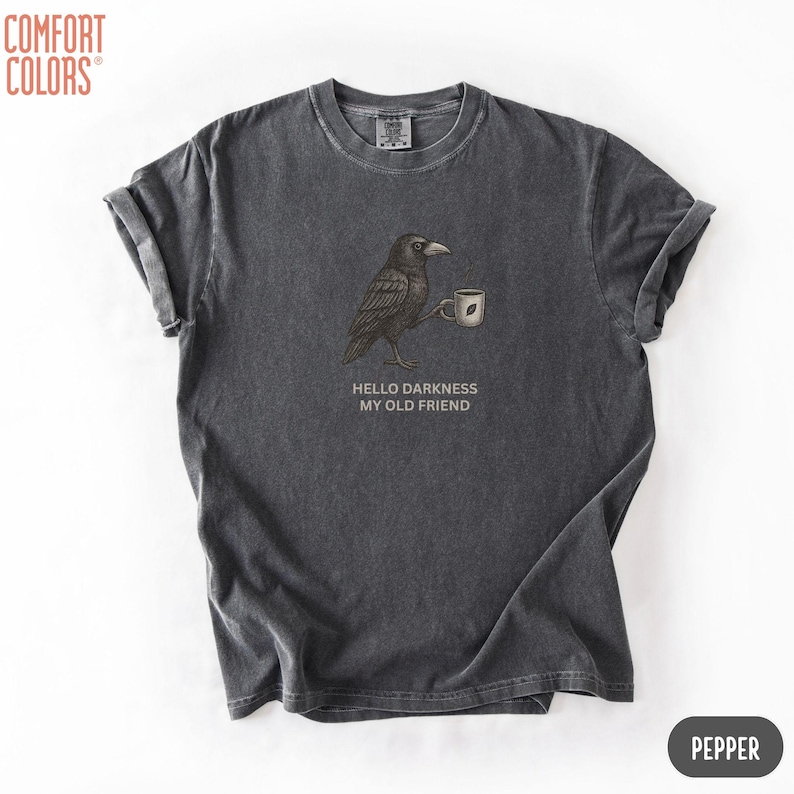 May include: A charcoal gray short-sleeve t-shirt featuring a graphic of a black crow holding a small white mug. Below the crow, the text reads HELLO DARKNESS MY OLD FRIEND. The shirt has a crew neckline and a vintage-style washed fabric finish.