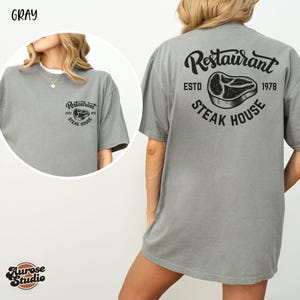 May include: Gray t-shirt with a restaurant logo on the front and back. The front logo says "Restaurant Steak House" and the back logo says "Restaurant ESTD 1978 Steak House".