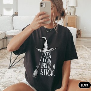 Puede incluir: Una camiseta negra de manga corta con un gr&aacute;fico blanco de un sombrero de bruja puntiagudo y una escoba. El texto impreso en blanco dice: YES I CAN DRIVE A STICK. La camiseta tiene cuello redondo y un corte holgado y c&oacute;modo.
