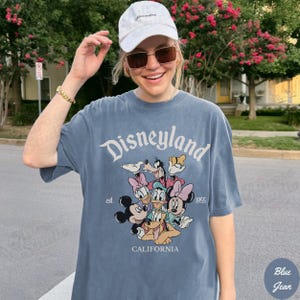 May include: Blue jean colored t-shirt with the word "Disneyland" in white script. Below the text is a graphic of Disney characters including Mickey and Minnie Mouse, Donald and Daisy Duck, and Goofy. The shirt also has the text "est. 1955" and "CALIFORNIA".