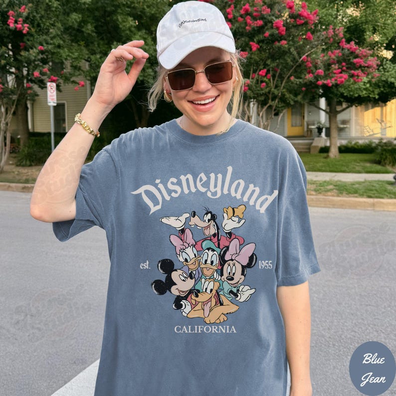 May include: Blue jean colored t-shirt with the word "Disneyland" in white script. Below the text is a graphic of Disney characters including Mickey and Minnie Mouse, Donald and Daisy Duck, and Goofy. The shirt also has the text "est. 1955" and "CALIFORNIA".