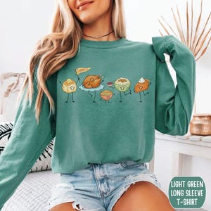 May include: A light green long-sleeve t-shirt featuring a graphic print of cartoon Thanksgiving food items including a dinner roll, a roasted turkey holding a flag that says THANKFUL, a bowl of stuffing, mashed potatoes, and a slice of pumpkin pie.