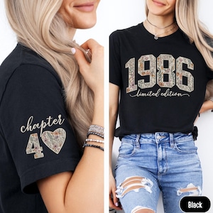 May include: Black t-shirt with the year "1986" in a floral design and the words "limited edition". The left sleeve has "chapter 40" with a floral heart. The shirt is worn with blue jeans.