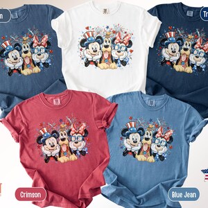 May include: Five Comfort Colors t-shirts in denim, white, true navy, crimson, and blue jean. Each shirt features a graphic of Mickey Mouse, Minnie Mouse, and Pluto in patriotic attire with fireworks.