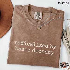 Pode incluir: Camiseta cor de café com a frase "radicalized by basic decency" em letras brancas. Um colar de prata com um pingente repousa sobre a camiseta. A camiseta está dobrada e um chapéu marrom é parcialmente visível.