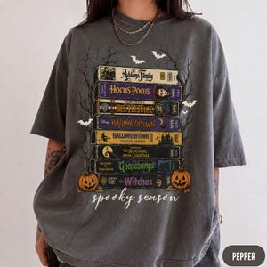 May include: A charcoal gray t-shirt featuring a graphic print of a stack of vintage VHS tapes with titles including The Addams Family, Hocus Pocus, Beetlejuice, Halloweentown, The Nightmare Before Christmas, Goosebumps, and The Witches, with spooky season text.