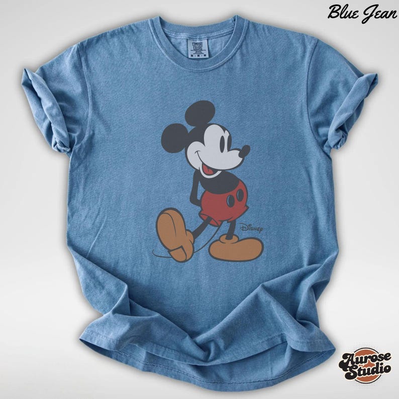 May include: Blue jean-colored t-shirt featuring a graphic of Mickey Mouse. The cartoon character is depicted in his classic outfit: red shorts, brown shoes, and black gloves. The shirt has rolled-up sleeves and the text "Blue Jean" is visible.