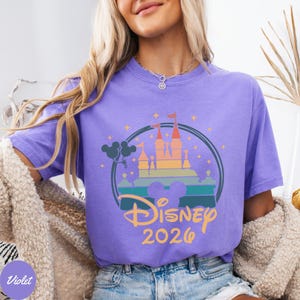 May include: A violet t-shirt featuring a colorful castle design within a circular frame. The design includes the text "Disney 2026" in a gold color, along with Mickey Mouse ears and star accents. The shirt is a solid violet color.