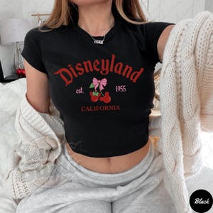 Puede incluir: Top corto negro con la palabra "Disneyland" en escritura roja, junto con "est. 1955" y "California". El dise&ntilde;o incluye un lazo rosa y cerezas. La camiseta es de manga corta.
