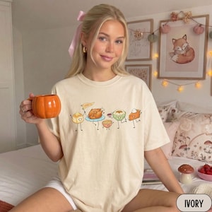 May include: An ivory t-shirt featuring a graphic print of cartoon Thanksgiving food items with arms and legs, including a turkey holding a flag that says Thankful, a bread roll, a bowl of mashed potatoes, a bowl of stuffing, and a slice of pumpkin pie.