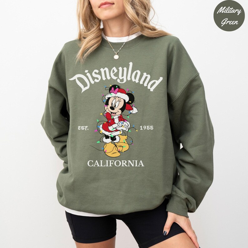 K&ouml;nnte beinhalten: Milit&auml;rgr&uuml;nes Disneyland-Sweatshirt mit einer Minnie Mouse-Grafik in einem Weihnachtsmannhut und -kleid, umwickelt von Lichterketten. Das Sweatshirt zeigt das Disneyland-Logo, "EST. 1955" und "CALIFORNIA".