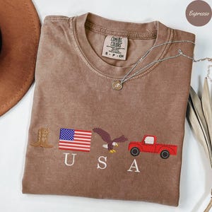 May include: Espresso-colored t-shirt with embroidered patriotic symbols: a cowboy boot, American flag, eagle, and red pickup truck. The word "USA" is embroidered below the symbols. A silver necklace with a pendant is draped across the shirt.