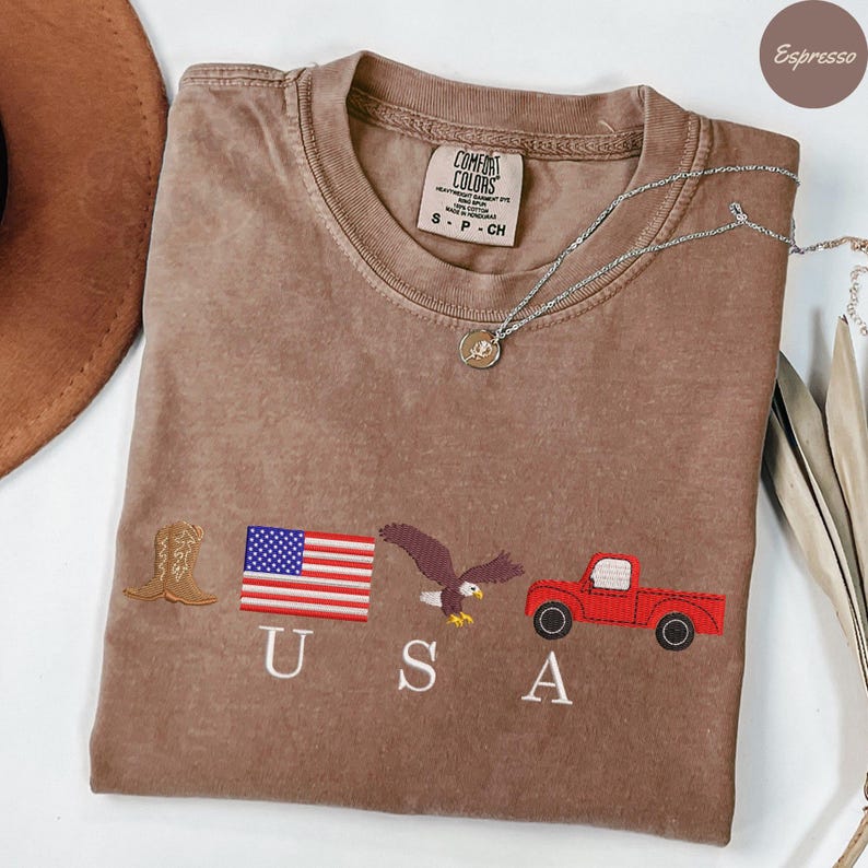 May include: Espresso-colored t-shirt with embroidered patriotic symbols: a cowboy boot, American flag, eagle, and red pickup truck. The word "USA" is embroidered below the symbols. A silver necklace with a pendant is draped across the shirt.