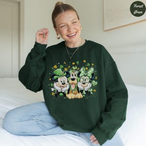 May include: A dark green sweatshirt featuring Mickey Mouse, Pluto, and Minnie Mouse in St. Patrick's Day attire. The graphic includes shamrocks and the words "Lucky Me". The sweatshirt is a comfortable, casual style.
